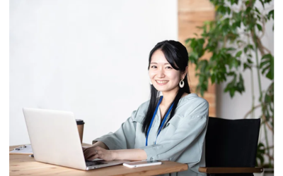 明るいオフィスで木製デスクに座り、ノートパソコンに向かって作業しながらカメラに笑顔を向ける、青いネックストラップを着用した若い女性。