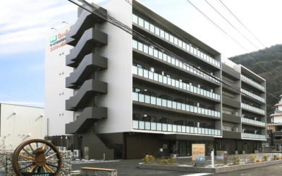 白とダークブラウンを基調としたマンション風の建物、「ココファン高松西宝町」の外観写真。手前には特徴的な大きな車輪のモニュメントが配置されている。